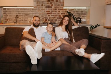 Baba, oğul ve anne evdeki kanepede televizyon izliyorlar. Mutlu aile evde akşamın tadını çıkarıyor. Uzun saçlı genç bir anne gülümsüyor..