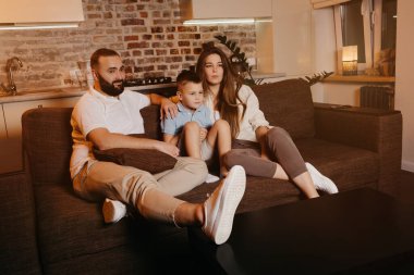 Sakallı baba, evlat ve uzun saçlı genç anne kanepede dikkatlice televizyon izliyorlar. Aile evde mutlu bir akşam geçiriyor..