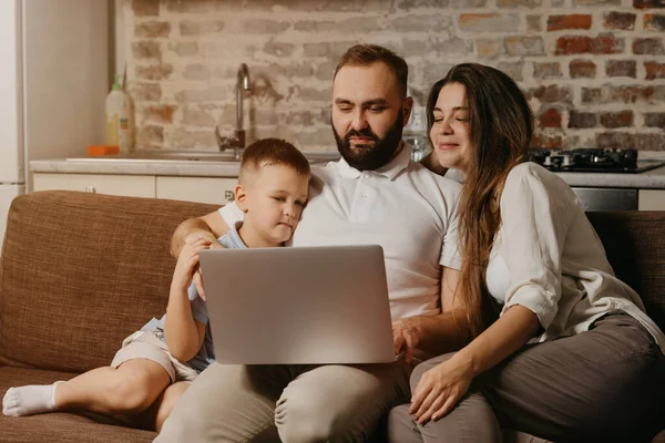 A father with a beard is working remotely on a laptop while his son and happy wife are staring at the screen. Dad is working online on a computer and hugging a child on the sofa at home in the evening