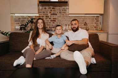 Baba, oğul ve anne evdeki kanepede TV ile izliyorlar. Aile evde mutlu bir akşamın tadını çıkarıyor. Uzun saçlı genç anne gülümsüyor..