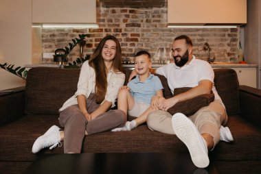 Baba, oğul ve anne evdeki kanepede televizyon izliyor ve gülüyorlar. Aile evde mutlu bir akşamın tadını çıkarıyor. Baba, oğluna ve karısına bakıyor..