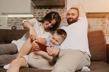Bir baba, bir oğul ve kanepede bir anne. Mutlu bir çocuk, akşam evde, gülümseyen anne ve babasının yanında akıllı telefondan maçın tadını çıkarıyor..