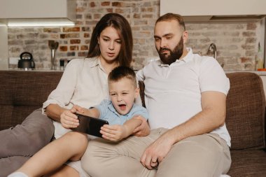 Bir baba, bir oğul ve bir anne kanepede video izliyorlar. Bir çocuk, akıllı telefondaki karikatür gösterisi sırasında akşam evde anne ve babasına esniyor..