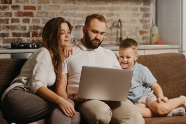 Sakallı bir baba, mutlu oğlu ve karısı ekrana bakarken uzaktan bilgisayarla çalışıyor. Babam bir bilgisayar üzerinde çalışıyor. Akrabaları arasında. Akşamları evdeki kanepede.