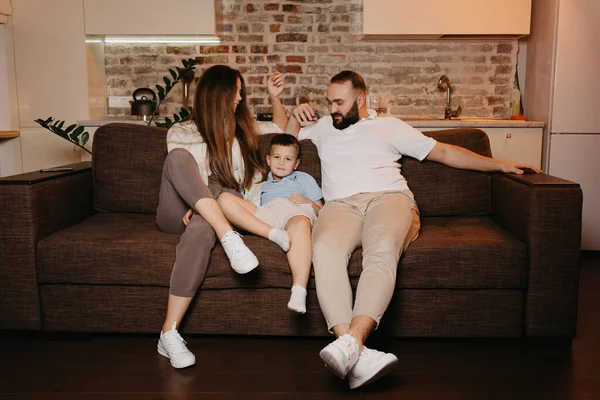 Dad, son, and mom are watching boring TV programs on the sofa in the apartment. The parents are staring at their boy in the evening. Relatives at home. A young mother with long hair is smiling.