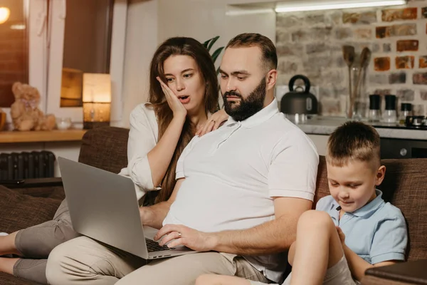 A serious father with a beard is working remotely on a laptop near his son while a wife is surprised at home. A family on the sofa in the evening. Dad is working online on a computer between relatives