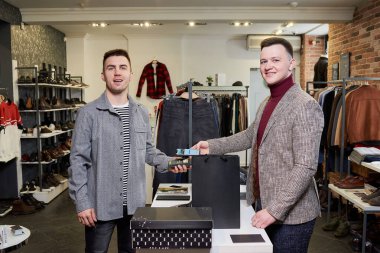 Bir adam akıllı telefonu kullanarak kablosuz NFC teknolojisiyle alışveriş yaparken poz veriyor. Gülümseyen bir tezgahtar bir butikte müşteriye ücretsiz ödeme yapmak için bir terminali soyuyor.