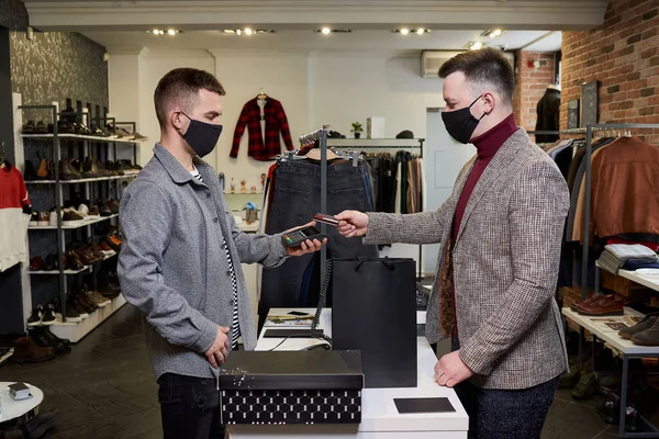 Man in a face mask is using a debit card o pay by wireless NFC technology for purchases in store. A shop assistant in mask is holding out a terminal for contactless paying to a customer in a boutique