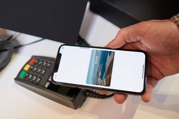 A close-up hand of a male customer who is holding a smartphone while paying through the digital wallet using NFC technology for his purchase in a store.
