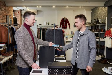 Bir adam akıllı telefonu kullanarak kablosuz NFC teknolojisiyle bir giyim mağazasından alışveriş yaparken poz veriyor. Gülümseyen bir tezgahtar müşteriye ücretsiz ödeme yapmak için bir terminal tutuyor.