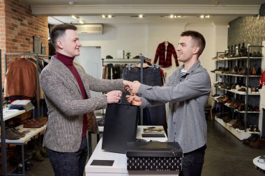 Mutlu bir adam alışverişini yapıyor ve giyim mağazasında gülümseyen bir tezgahtarla el sıkışıyor..