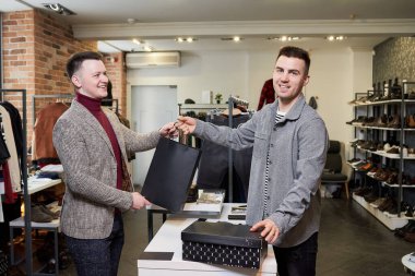 Mutlu bir adam alışverişini bir giyim mağazasından yapıyor. Gülümseyen bir erkek tezgahtar içinde kıyafet olan bir kese kâğıdı ve şık bir mağaza müşterisine ayakkabı kutusu veriyor..