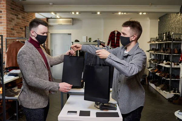 A man in a face mask to avoid the spread of coronavirus is taking his purchases from a seller in a clothing store. A male shop assistant is giving paper bags with clothes to a customer in a boutique.