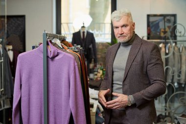 Gri saçlı olgun bir adam giyim mağazasından balıkçı yaka kazak seçerken poz veriyor. Butikte yün takım elbiseli bir erkek müşteri..