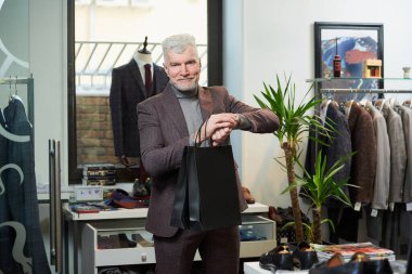 Gri saçlı, sportif fiziği olan mutlu, olgun bir adam bir giyim mağazasında elinde iki siyah kese kâğıdıyla kol saatine bakıyor. Sakallı bir erkek müşteri butikte yün takım elbise giyer.