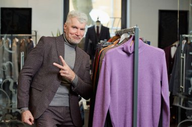 Gri saçlı olgun bir adam giyim mağazasında zafer işareti gösteriyor. Butikte yün takım elbiseli mutlu bir erkek müşteri..