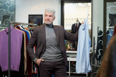 Gri saçlı olgun bir adam bir giyim mağazasında gömlek gösteriyor. Sakallı bir erkek müşteri butikte yün takım elbise giyer..
