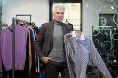 Gri saçlı olgun bir adam elbise mağazasında poz veriyor ve gömlek gösteriyor. Sakallı bir erkek müşteri butikte yün takım elbise giyer..