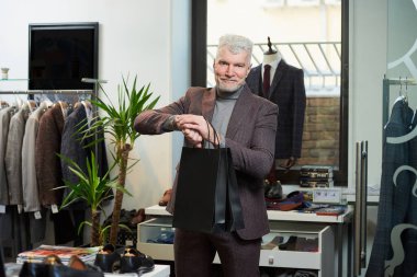 Gri saçlı, sportif fiziği olan mutlu, olgun bir adam bir giyim mağazasında elinde iki siyah kese kâğıdıyla kol saatine bakıyor. Sakallı bir erkek müşteri butikte yün takım elbise giyer.