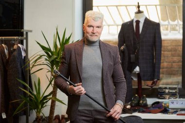 Gri saçlı ve spor fiziği olan gülümseyen olgun bir adam elbise mağazasında iki eli olan lifli bir karbon baston tutuyor. Sakallı bir erkek müşteri butikte takım elbise giyer..