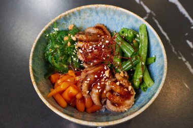 Üzerinde ahtapot, Wakame yosunu, havuç, fıstık, susam, siyah taş bir masada beyaz pirinç olan bir dürtme kabının üst görüntüsü..