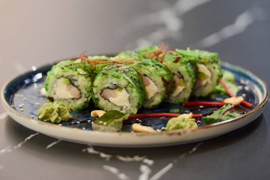 Chuka Wakame deniz yosunu, Philadelphia peyniri ve mavi seramik tabakta soslu karides, kaju ve wasabi ile siyah taş bir masada çekilmiş suşi rulolarının yakın plan fotoğrafı..
