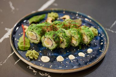 Chuka Wakame deniz yosunu, Philadelphia peyniri ve mavi seramik tabakta soslu karides, kaju ve wasabi ile siyah taş bir masada çekilmiş suşi rulolarının yakın plan fotoğrafı..