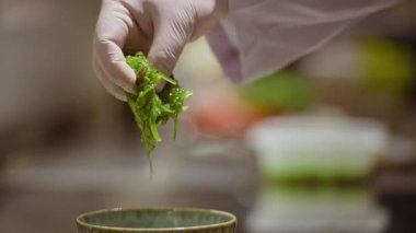 Chuka Wakame 'yi mutfaktaki seramik kaseye atan tek kullanımlık eldivenli bir erkek aşçının yakın çekim videosu..