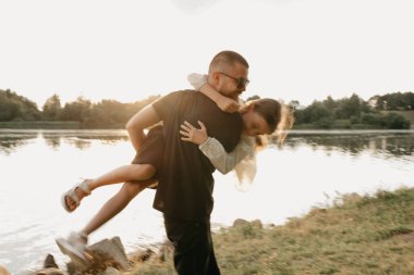 Gölün kıyısında kızının kollarında dolanan sakallı genç bir babanın bulanık fotoğrafı. Günbatımında tatilde olan tek ebeveynli şık bir aile..