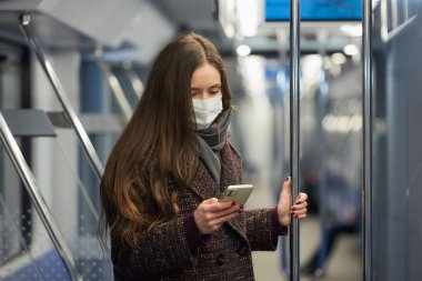Coronavirus 'un yayılmasını önlemek için tıbbi maske takan bir kadın modern bir metro vagonunda ayakta duruyor ve akıllı telefon kullanıyor. Cerrahi maskeli bir kız trende cep telefonuyla haberlere çıkıyor..