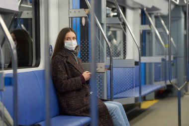 Coronavirus 'un yayılmasını önlemek için tıbbi maske takan bir kadın modern bir metro vagonunda yan yana oturuyor. COVID-19 'a karşı cerrahi maske takan bir kız metro trenine biniyor..