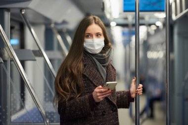 Coronavirus 'un yayılmasını önlemek için maske takan kadın modern bir metro vagonunda ayakta duruyor ve yan tarafa bakıyor. COVID-19 'a karşı cerrahi maske takan bir kız metro trenine biniyor.