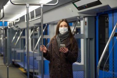 Coronavirus 'un yayılmasını önlemek için tıbbi maskeli bir kadın modern bir metro vagonunda ayakta duruyor ve elinde bir akıllı telefon tutuyor. COVID-19 'a karşı ameliyat maskeli bir kız metroya biniyor..