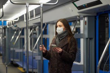 Coronavirus 'un yayılmasını önlemek için tıbbi maske takan bir kadın modern bir metro vagonunda duruyor ve elinde telefon tutuyor. COVID-19 'a karşı cerrahi maske takan bir kız metro trenine biniyor..