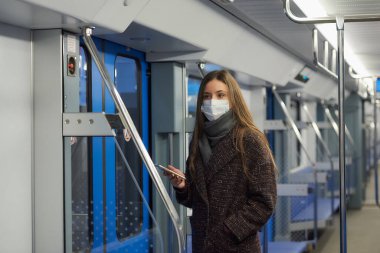 Coronavirus 'un yayılmasını önlemek için tıbbi maske takan bir kadın kapıların yanında duruyor ve boş bir metro vagonunda yan tarafa bakıyor. Cerrahi maskeli bir kız metro trenine biniyor..