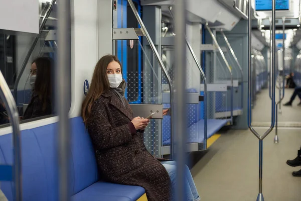 A woman in a medical face mask to avoid the spread of coronavirus is sitting holding a smartphone in a modern subway car. A girl in a surgical mask against COVID-19 is taking a ride on a metro train.