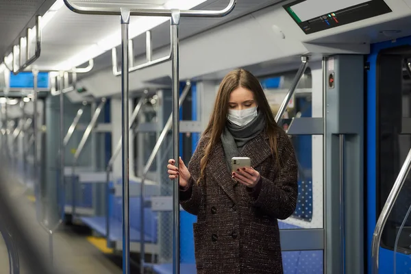 A woman in a medical face mask to avoid the spread of coronavirus is standing and using a smartphone in an empty subway car. A girl in a surgical mask is scrolling news on her cellphone on a train.