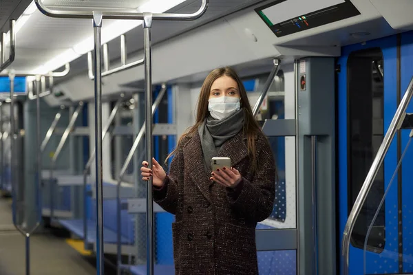 Woman in a medical face mask to avoid the spread of coronavirus is standing and holding a smartphone in a modern subway car. Girl in a surgical mask against COVID-19 is taking a ride on a metro train.