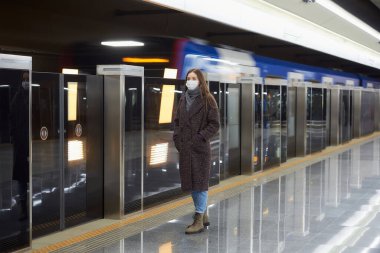 Metroda bir trenin gelmesini bekleyen Coronavirus 'un yayılmasını önlemek için tıbbi maske takan bir kadının tam boy fotoğrafı. Maskeli bir kız sosyal mesafeyi koruyor..