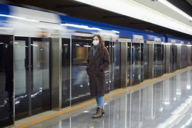 Metroda modern bir trenin gelmesini bekleyen Coronavirus 'un yayılmasını önlemek için tıbbi maske takan bir kadının tam boy fotoğrafı. Cerrahi maskeli kız sosyal mesafeyi koruyor..