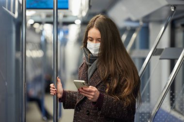 Coronavirus 'un yayılmasını önlemek için tıbbi maske takan bir kadın modern bir metro vagonunda ayakta duruyor ve akıllı telefon kullanıyor. Cerrahi maskeli bir kız trende cep telefonuyla haberlere çıkıyor..