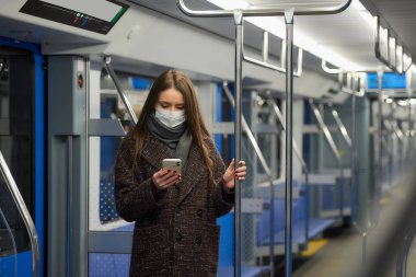 Coronavirus 'un yayılmasını önlemek için tıbbi maske takan bir kadın boş bir metro vagonunda akıllı telefon kullanıyor. Cerrahi maskeli bir kız trende cep telefonuyla haberlere çıkıyor..