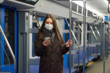 Coronavirus 'un yayılmasını önlemek için tıbbi maske takan bir kadın boş bir metro vagonunda akıllı telefon kullanıyor. Cerrahi maskeli bir kız trende cep telefonuyla haberlere çıkıyor..