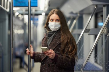 Coronavirus 'un yayılmasını önlemek için maske takan kadın modern bir metro vagonunda ayakta duruyor ve yan tarafa bakıyor. COVID-19 'a karşı cerrahi maske takan bir kız metro trenine biniyor.