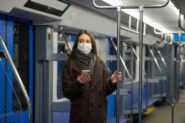 Coronavirus 'un yayılmasını önlemek için tıbbi maskeli bir kadın modern bir metro vagonunda ayakta duruyor ve elinde bir akıllı telefon tutuyor. COVID-19 'a karşı ameliyat maskeli bir kız metroya biniyor..