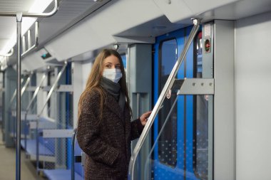 Coronavirus 'un yayılmasını önlemek için tıbbi maske takan bir kadın kapıların yanında duruyor ve modern bir metro vagonunun kenarına bakıyor. Cerrahi maskeli bir kız metro trenine biniyor..