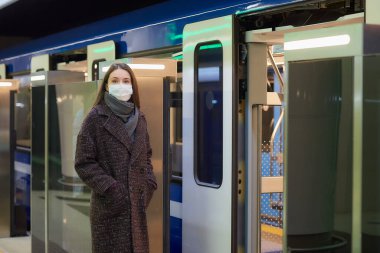 Koronavirüsün yayılmasını önlemek için tıbbi maske takan bir kadın modern metro vagonunun yanında bekliyor. Cerrahi maskeli bir kız metro istasyonunda sosyal mesafeyi koruyor..