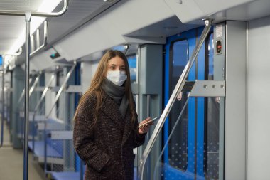 Coronavirus 'un yayılmasını önlemek için tıbbi maske takan bir kadın kapıların yanında duruyor ve boş bir metro vagonunda yan tarafa bakıyor. Cerrahi maskeli bir kız metro trenine biniyor..