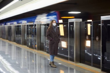 Metroda bir trenin gelmesini bekleyen Coronavirus 'un yayılmasını önlemek için tıbbi maske takan bir kadının tam boy fotoğrafı. Maskeli bir kız sosyal mesafeyi koruyor..