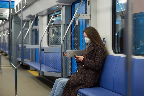 A woman in a medical face mask to avoid the spread of coronavirus is sitting and using a smartphone in a modern subway car. A girl in a surgical mask is scrolling news on her cellphone on a train.